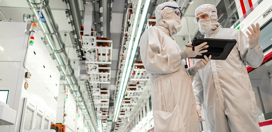 Two co-workers in a fab plant, in clean room suits, discussing a laptop screen.