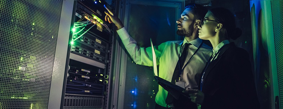 Two people in dark server room pointing at equipment